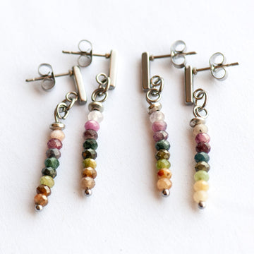 The Spectrum Spark – Tourmaline Rainbow Earrings by Fierce Lynx Designs feature four dangling earrings with silver-plated studs and faceted tourmaline beads in green, pink, yellow, and brown—perfect for rainbow gemstone lovers.