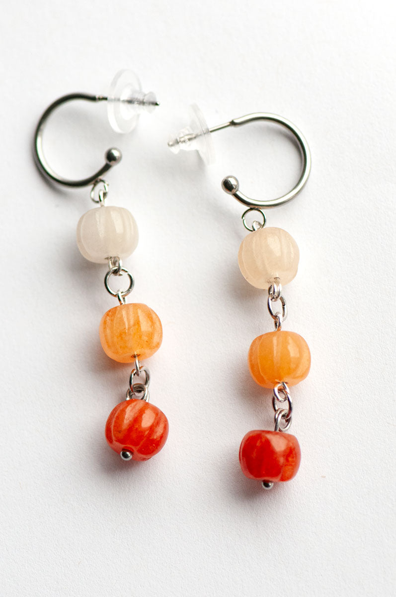 Introducing the Pumpkin Spice Earrings by Fierce Lynx Designs: a pair of silver hoop earrings, each adorned with three round beads—white, orange aventurine, and red—arranged from top to bottom. Perfect as pumpkin earrings or fall jewelry, these stylish hoops are beautifully displayed on a white background.