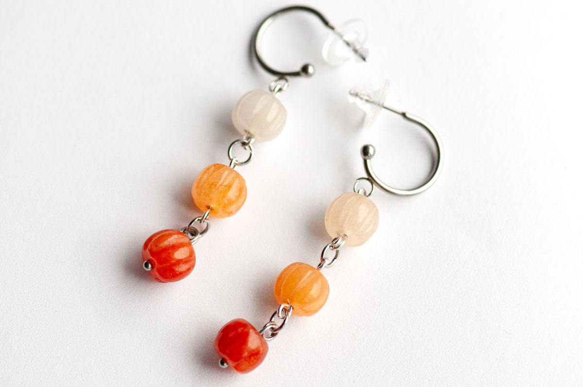 The Pumpkin Spice Earrings by Fierce Lynx Designs feature two silver dangle earrings, each adorned with three round beads in a gradient of fall-inspired colors—rich red at the bottom, orange aventurine in the center, and pale beige at the top. Evoking a subtle nod to pumpkin motifs, these earrings are perfect as autumn jewelry. Shown on a white background.