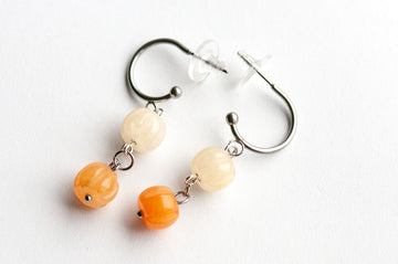 The Pumpkin Spice Earrings by Fierce Lynx Designs are a pair of metal clip-on earrings featuring two round beads on each—one cream-colored and one orange aventurine—evoking the warm, autumnal tones of fall jewelry, beautifully displayed against a white background.