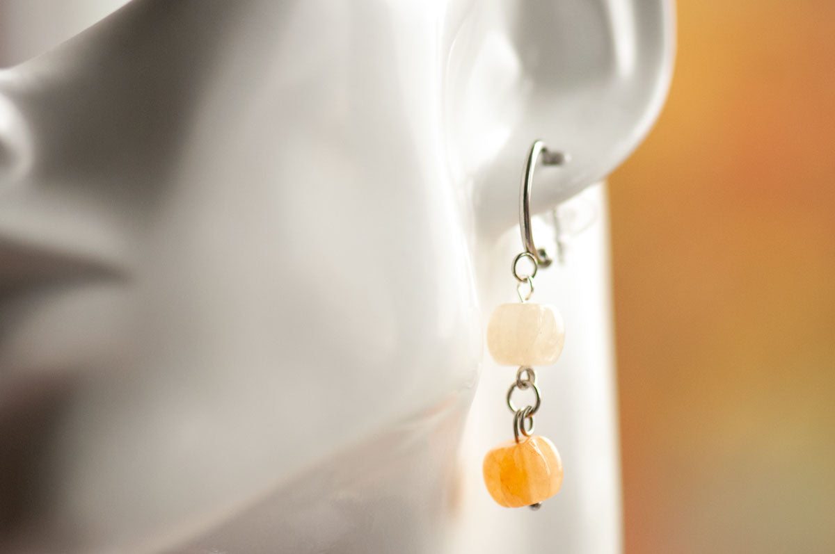 A close-up shot highlights an ear adorned with Fierce Lynx Designs’ Pumpkin Spice Earrings—dangling with two square beads, one white and one orange aventurine, connected by metal links. The softly blurred, warm-toned background perfectly complements this unique fall jewelry piece.