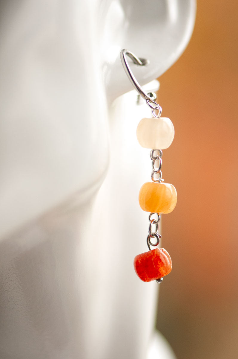 Close-up of a person wearing Fierce Lynx Designs’ Pumpkin Spice Earrings—dangling with three stacked beads in white, orange aventurine, and red, connected by metal links. Perfect for fall jewelry lovers; the blurred background features warm tones.