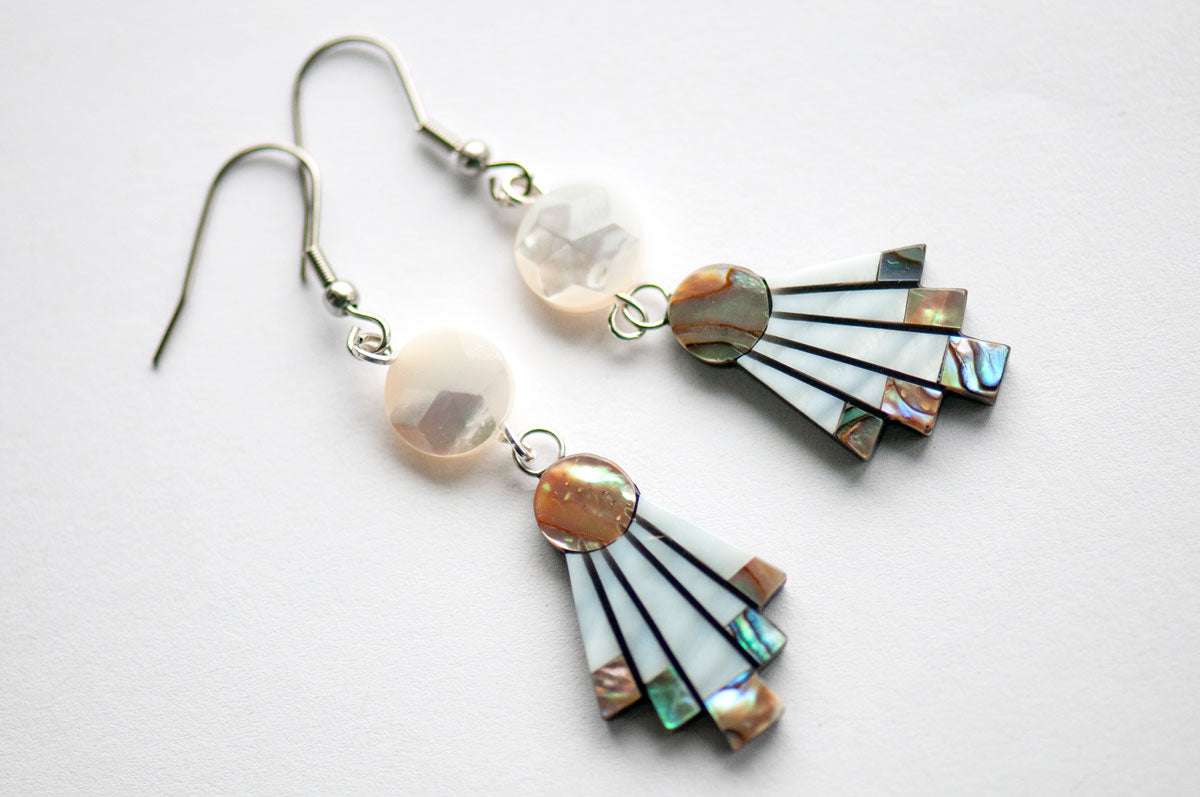 The Mother of Pearl Flare Earrings by Fierce Lynx Designs are a pair of statement dangle earrings featuring round white beads and fan-shaped pendants with iridescent and white segments, displayed on a white background.