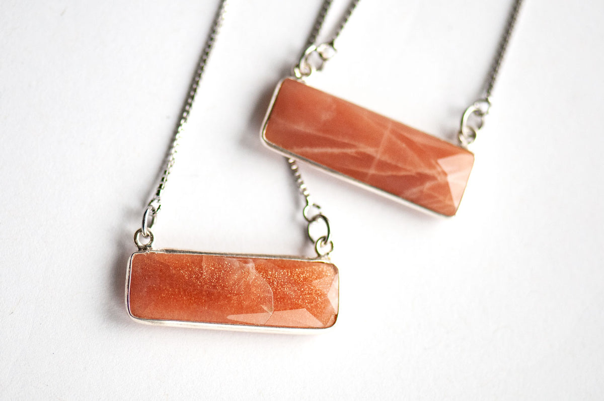 Angled close-up of two faceted peach bar pendants in silver bezels, showing glittering flecks in one and moonstone sheen lines in the other.